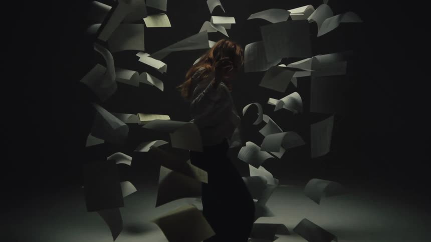A woman dances gracefully in slow motion amidst a chaotic whirlwind of floating papers in a dark, dramatically lit room