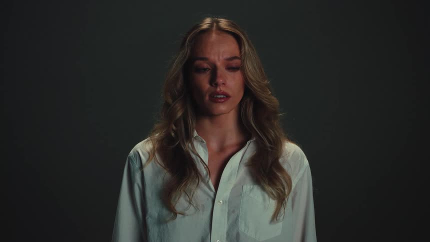 A heartbroken blonde woman cries a single tear in slow motion against a dark background