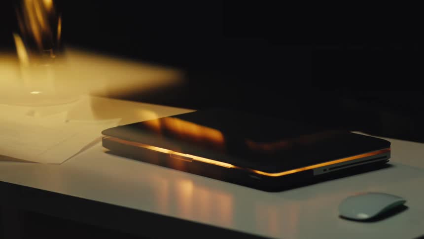 A sleek black and gold laptop rests on a desk, its surface reflecting a warm, flickering light in a dark room