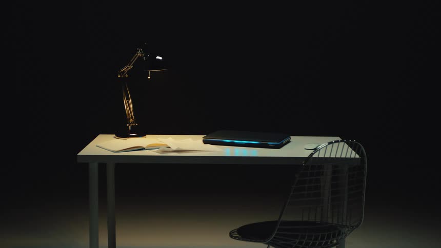 A modern desk with a closed laptop glowing with a blue light sits under a lamp in a dark, quiet room