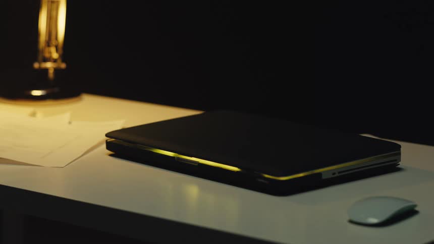 A closed laptop glows with pulsating colored light on a desk in a dark room