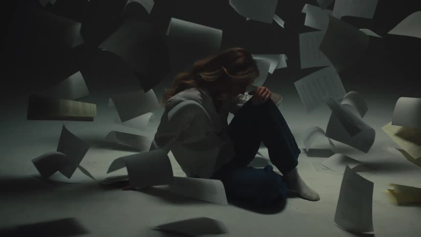 A stressed and overwhelmed woman sits alone in a dark room while sheets of paper float around her in slow motion