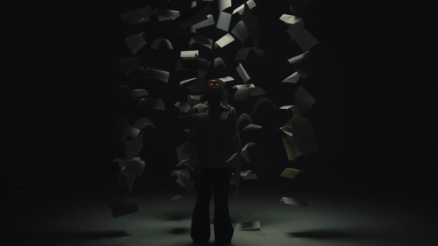 A woman stands in a spotlight as papers fall down around her in slow motion in a dark, empty room