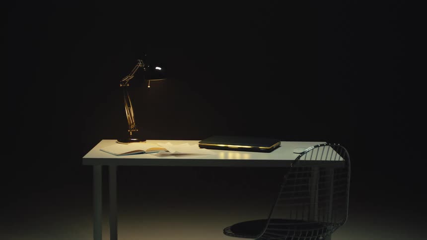 Slow dolly out from a modern, minimalist desk and chair illuminated by a single lamp in the dark