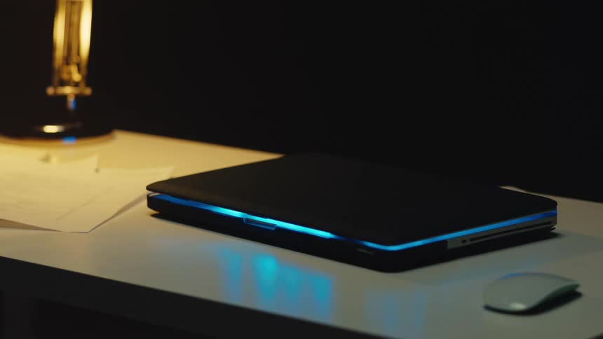 A modern laptop with a pulsing blue light in sleep mode rests on a desk in a dark room