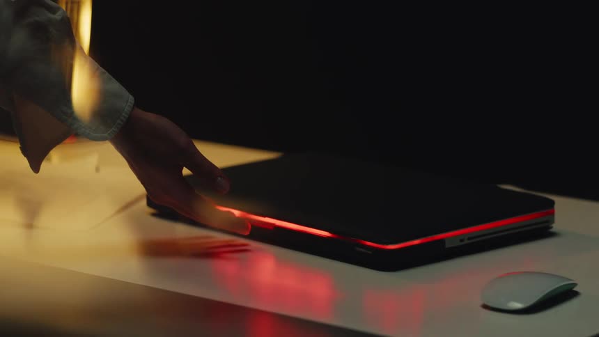 A hand slowly opens a modern laptop with a glowing red LED light strip on a white desk in a dark room