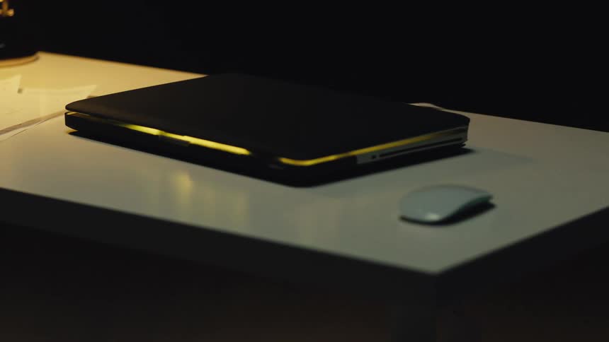 A closed laptop on a desk glows and flickers with an inner light in a dark room