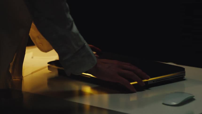 Ethereal light leaks from a laptop as a person closes the lid in a dark, moody setting