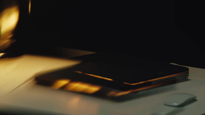 Flickering warm light from a lamp reflects off a closed laptop on a desk in a dark room
