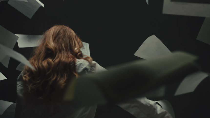 A stressed woman is surrounded by papers flying through the air in slow motion, feeling overwhelmed by information and work