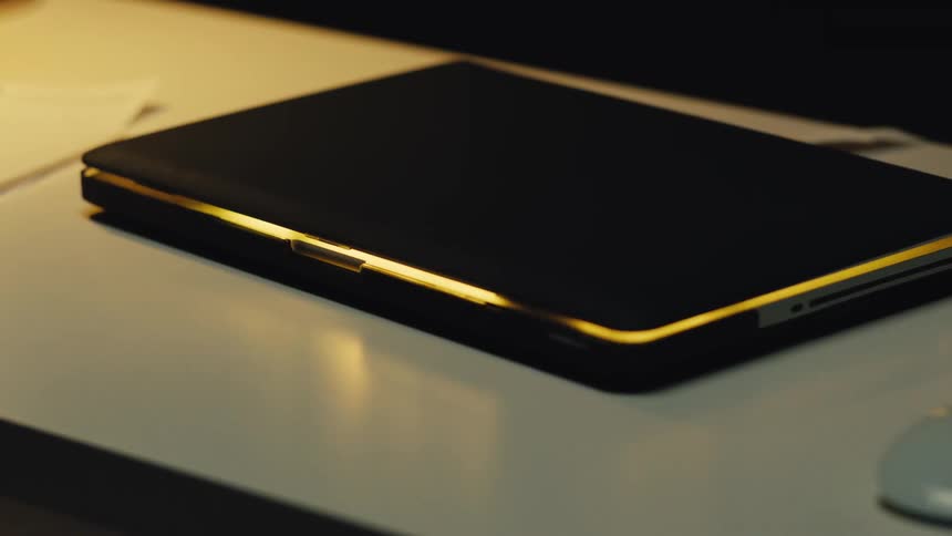 Pulsing light glows from within a closed laptop resting on a desk in a dimly lit room