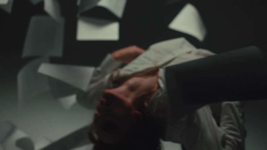 A woman in emotional distress contorts her body backward as a storm of paper flies around her in a dark room in slow motion