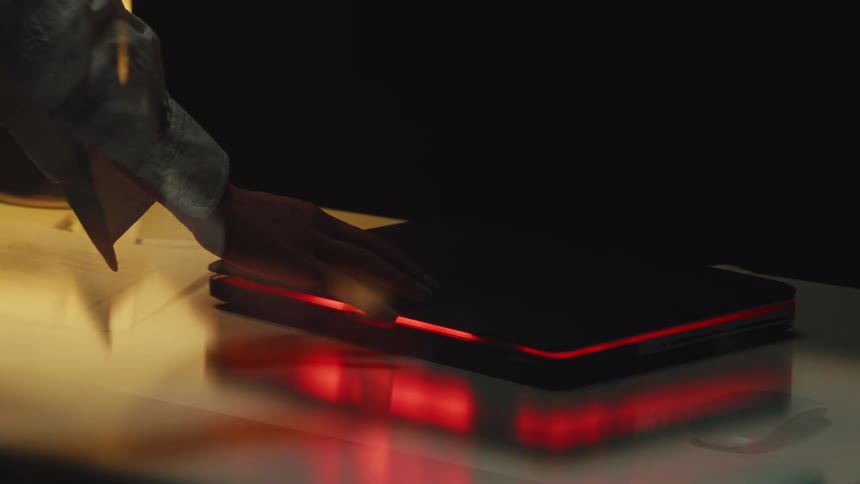 A person's hand slowly closes a modern laptop with a glowing red light strip on a desk in a dark room