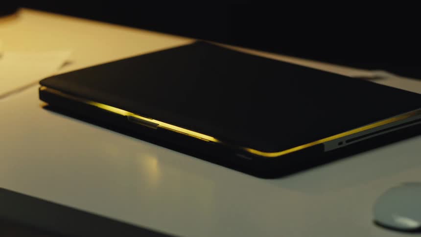 A laptop computer pulses with a mysterious golden light while closed on a desk in a dark room