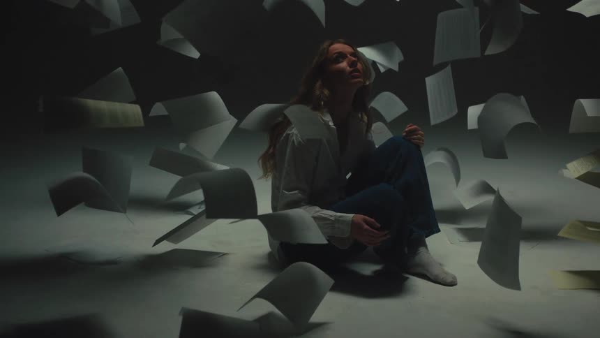 An overwhelmed woman sits on the floor in a dark room while papers and documents float around her in slow motion