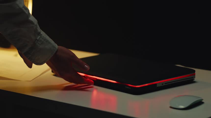 A person's hand controls the pulsating red light on a futuristic closed laptop using gestures in a dark room