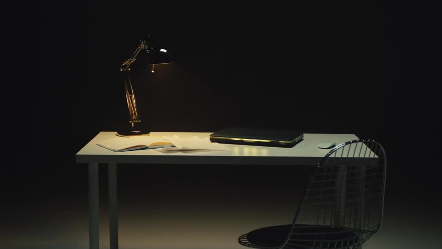 A single lamp illuminates an empty, modern desk with a laptop and notebook in a dark room