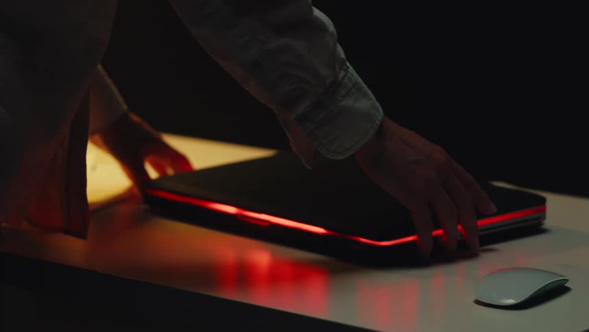 A person's hands nervously tap and slide across a closed, glowing laptop in a dark room
