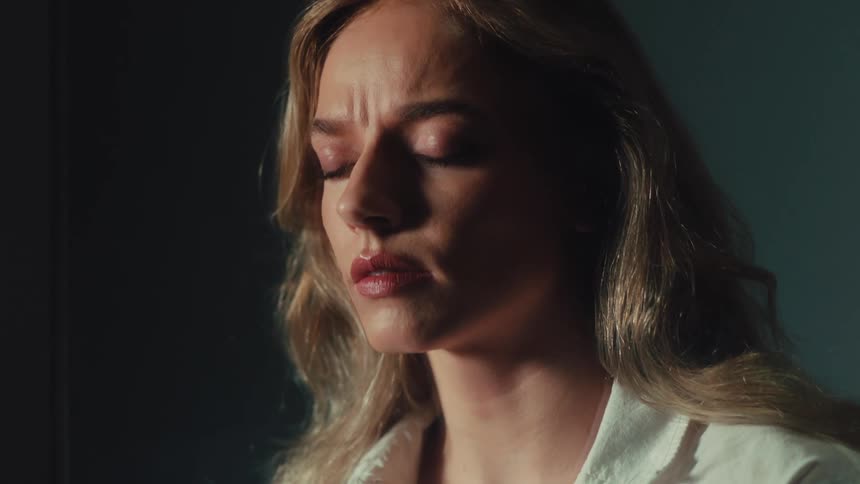 A close-up of a distraught and sad woman with her eyes closed, feeling worried and stressed in a dramatically lit room