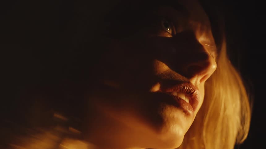A dramatic low-angle close-up of a woman's face illuminated by a warm, flickering light source in the dark