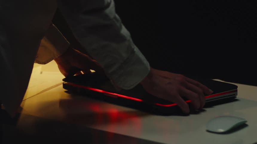 Close up shot of person's hands closing a sleek laptop with a glowing red light in a dimly lit office