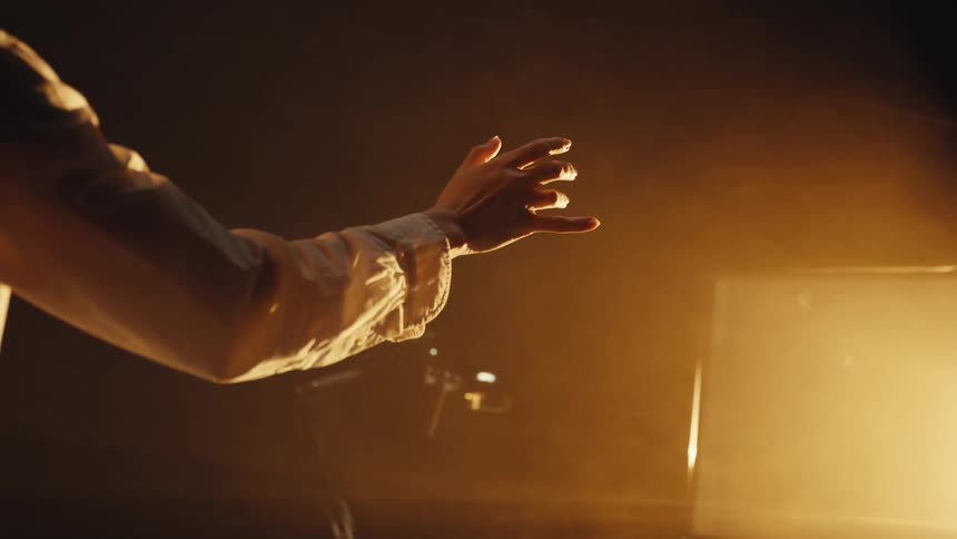 A person's hand moves gracefully in slow motion through a warm, smoky spotlight in a dark room
