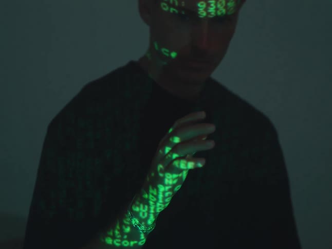 A man stands in darkness as green lines of code are projected onto him, he then slowly raises his hand toward the camera