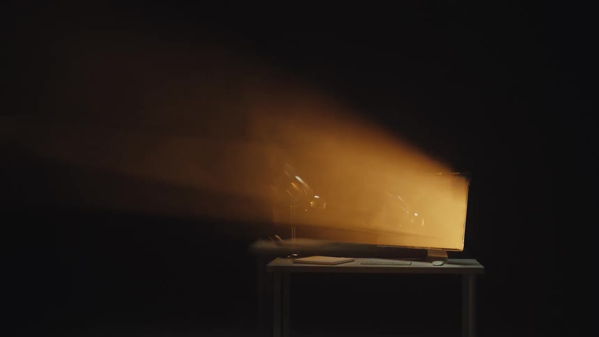 Dramatic time-lapse of a computer desk with a brightly lit monitor creating light beams in a dark, smoky room