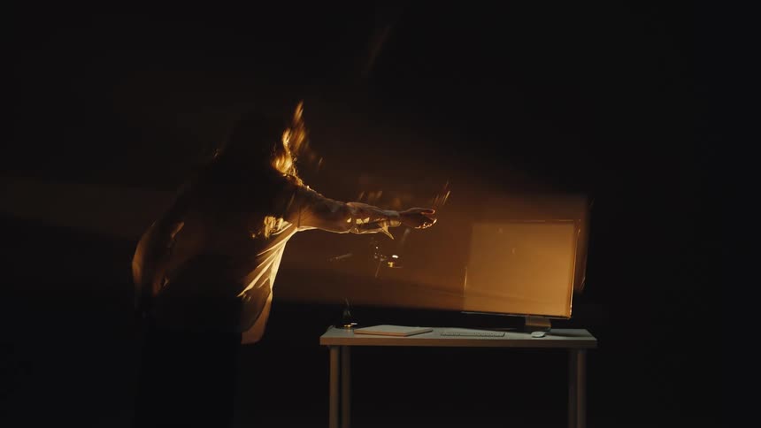 An artistic long exposure shot captures a stressed person shattering a wine glass in front of a glowing computer monitor in a dark room
