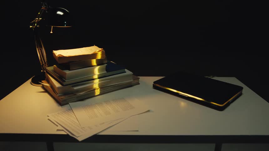A shimmering golden light scans across a stack of old books and a laptop illuminated by a single desk lamp in a dark, mysterious room