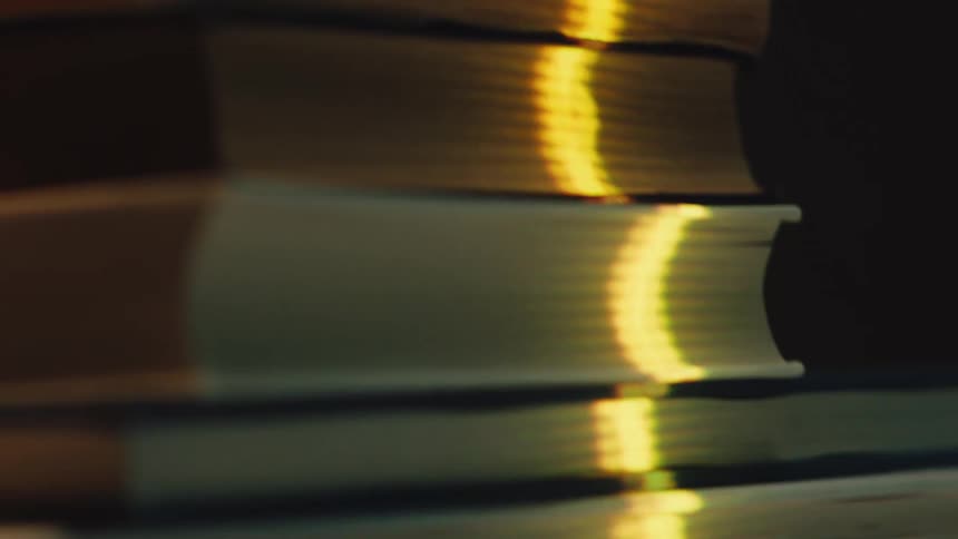 A slow-motion close-up reveals a stack of old books illuminated by a single, warm ray of sunlight against a dark background