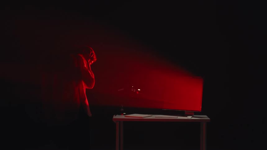 A disturbed person in a hoodie reacts angrily to what they see on a computer screen, illuminated by its menacing red glow in a dark room