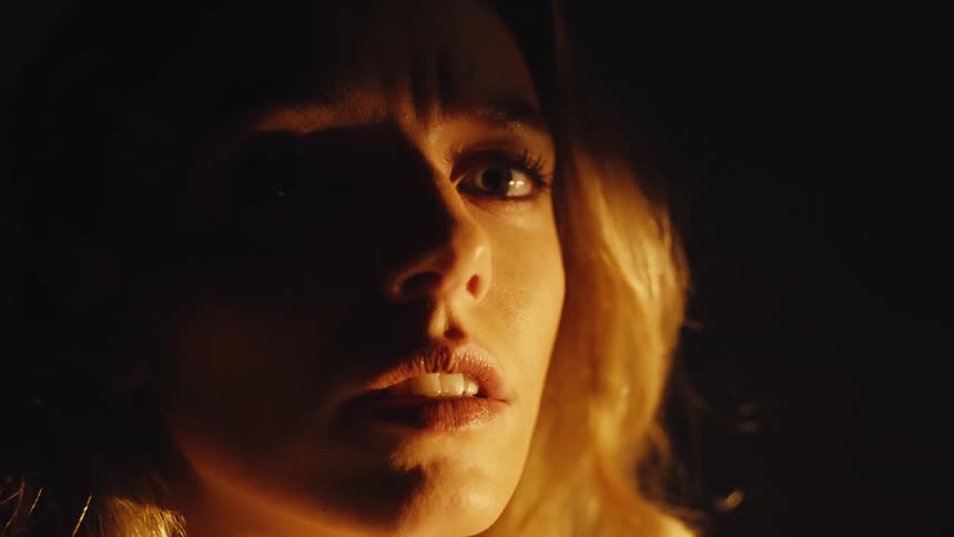 A close-up of a woman's worried face illuminated by warm light in the dark as her expression slowly changes to fear