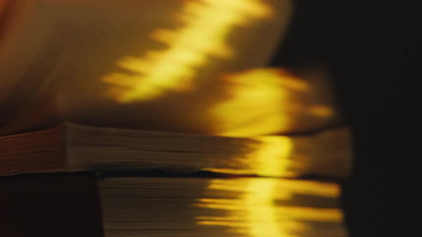 A shimmering golden light reflects on the pages of a book fluttering in the wind, creating an abstract and artistic visual