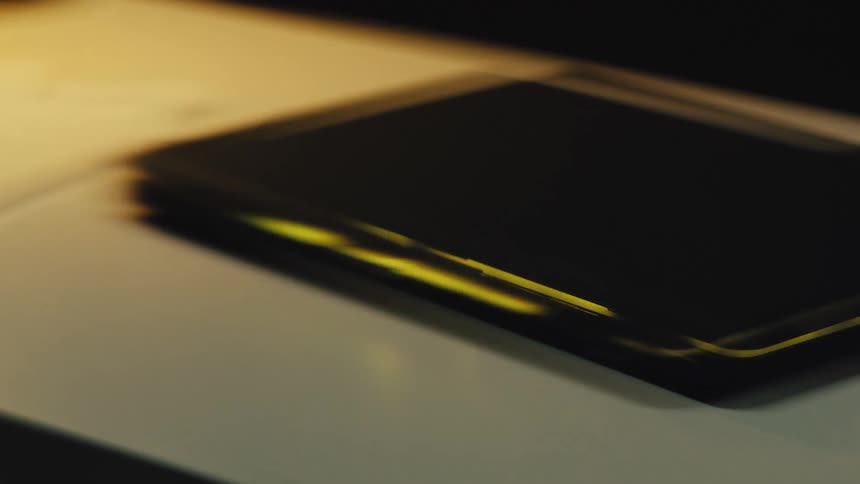 A closed laptop on a white desk is illuminated by the flickering warm light of a nearby lamp, creating abstract reflections