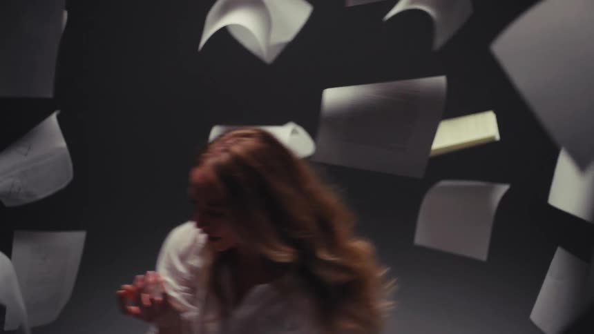 A woman in a white shirt moves in slow motion while papers fall all around her in a dark, dramatic setting
