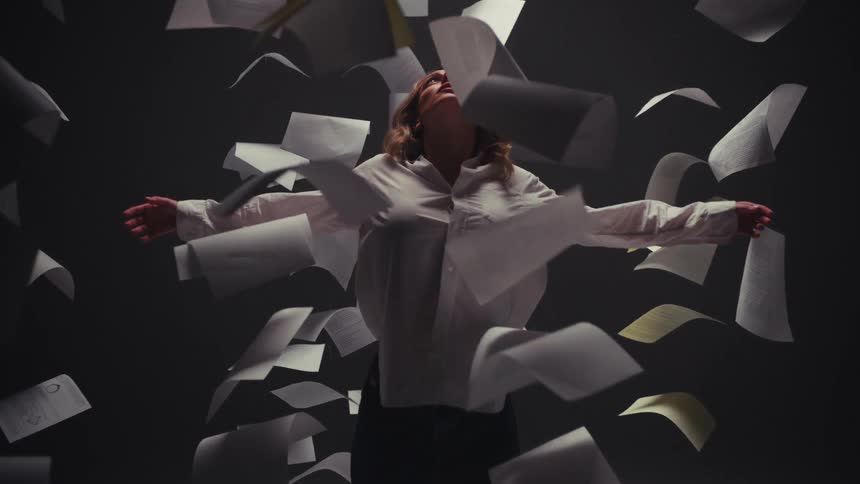 A woman stands with her arms outstretched in a dramatic, dark room as sheets of paper swirl around her in slow motion