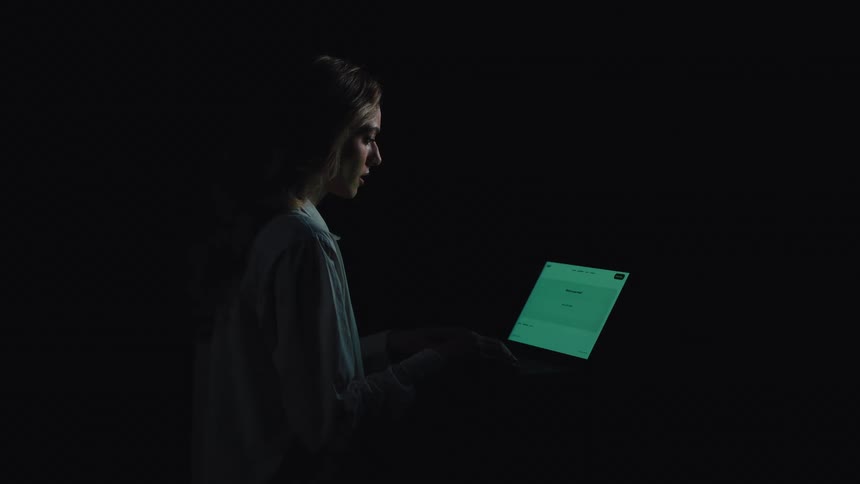 Side view of a woman sitting in a dark room, her face illuminated only by the glowing screen of her laptop as she types