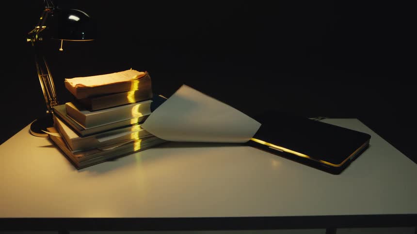 A strong wind blows through a dark room, turning the pages of an old book and scattering papers across a desk in slow motion