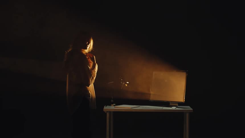 A woman illuminated by a single spotlight prepares her microphone and headphones for a recording session in a dark studio