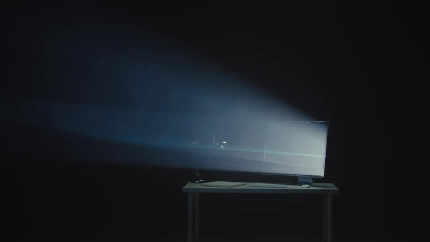 Bright light beams emanate from a computer monitor on a desk in a dark, hazy room