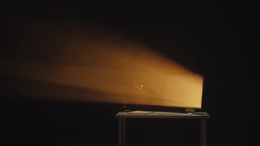 A computer monitor on a desk emits a powerful, hazy beam of golden light in a dark, empty room