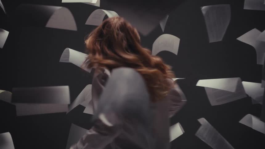 An expressive woman dances in slow motion, surrounded by a whirlwind of falling documents in a dark, moody studio