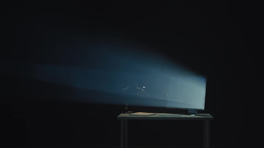 A computer monitor on a desk glows brightly, casting a volumetric beam of light through a dark, hazy room