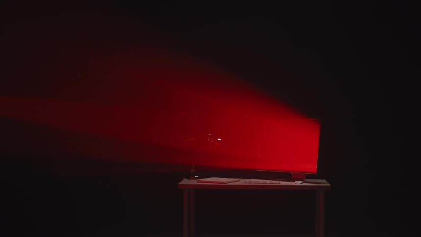 A red beam of light from a projector illuminates a desktop computer on a desk in a dark, hazy room
