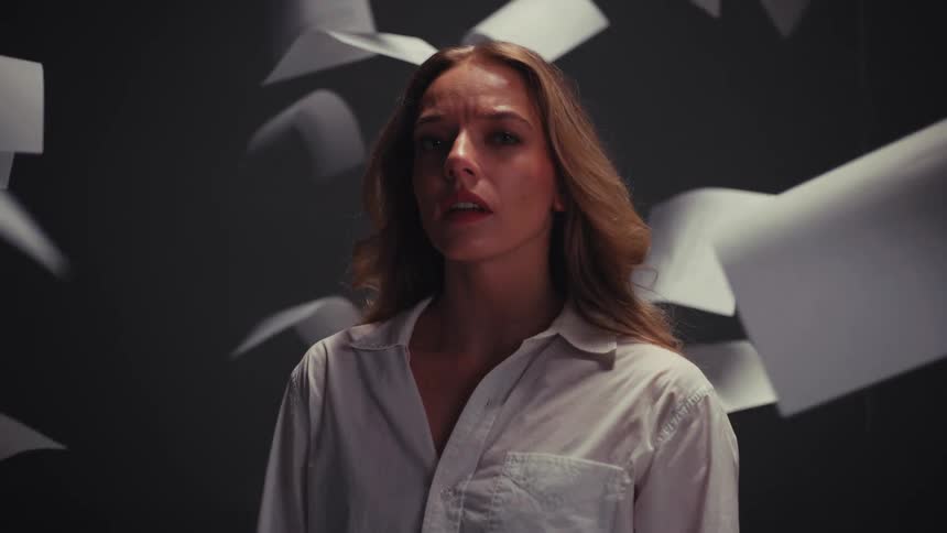A stressed and overwhelmed woman stands still while sheets of paper float and fall around her in slow motion