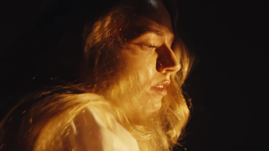 A distressed blonde woman writhes in slow motion, her anguished face illuminated by warm, dramatic light in the darkness