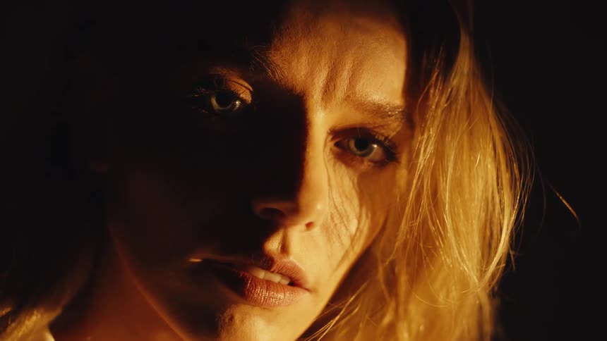An extreme close-up of a terrified woman's face lit by a single warm light in a dark room as she stares in horror