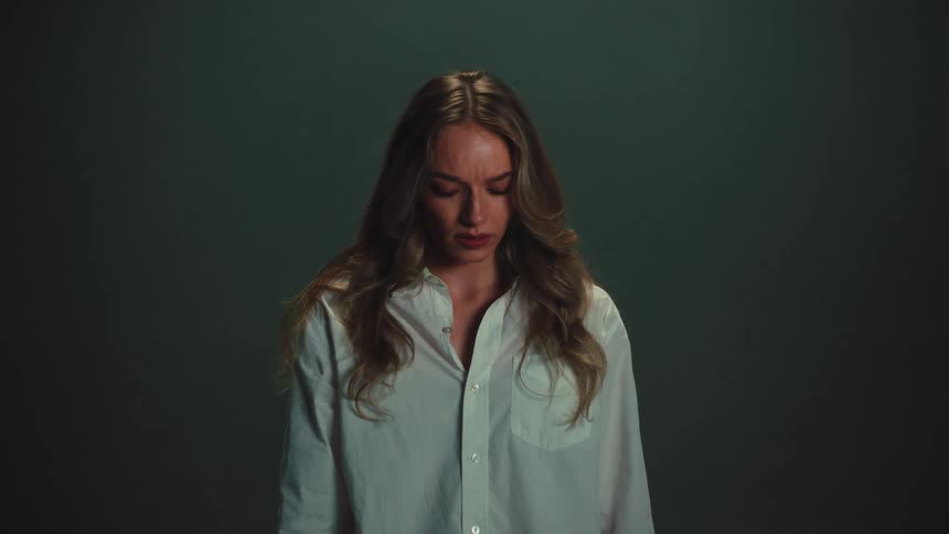 A sorrowful woman in a spotlight is showered with falling papers, her expression shifting from curiosity to deep despair