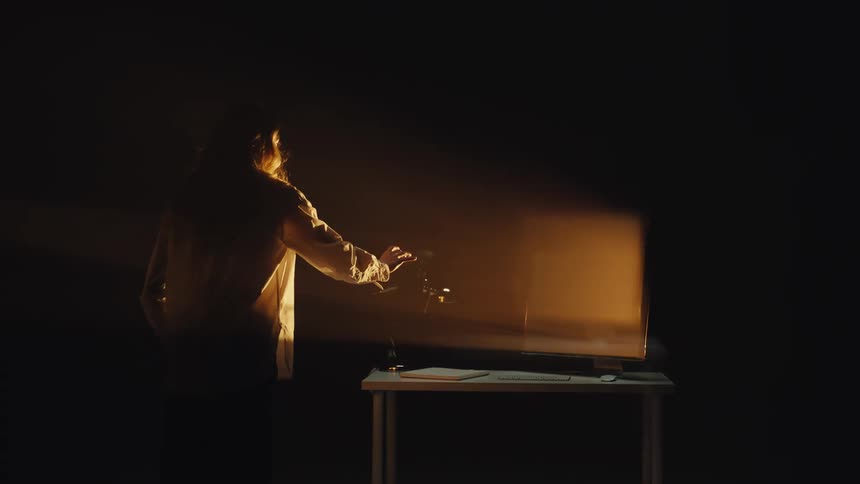 A long exposure shot captures a shadowy figure in a dark, moody room smashing a microphone on a desk in a fit of rage